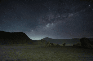 Night photography at Mount Bromo with a person standing beside the Bromo Tengger Semeru National Park entrance sign under a bright Milky Way galaxy sky.