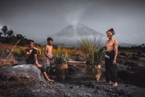 Concept photography of local villagers in Pronojiwo, Lumajang, with Mount Semeru volcano in the background. An elderly man and two boys interact joyfully near woven baskets filled with harvested grass. Captured in natural light, the scene highlights traditional Indonesian rural life, culture, and breathtaking volcanic landscape.