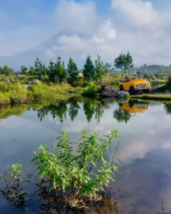 Yellow jeep parked beside a clear water pond with Mount Semeru volcano in the background, surrounded by green trees and blue sky. Adventure travel photography in Pronojiwo, Lumajang, highlighting nature, reflection, and outdoor exploration in East Java, Indonesia.