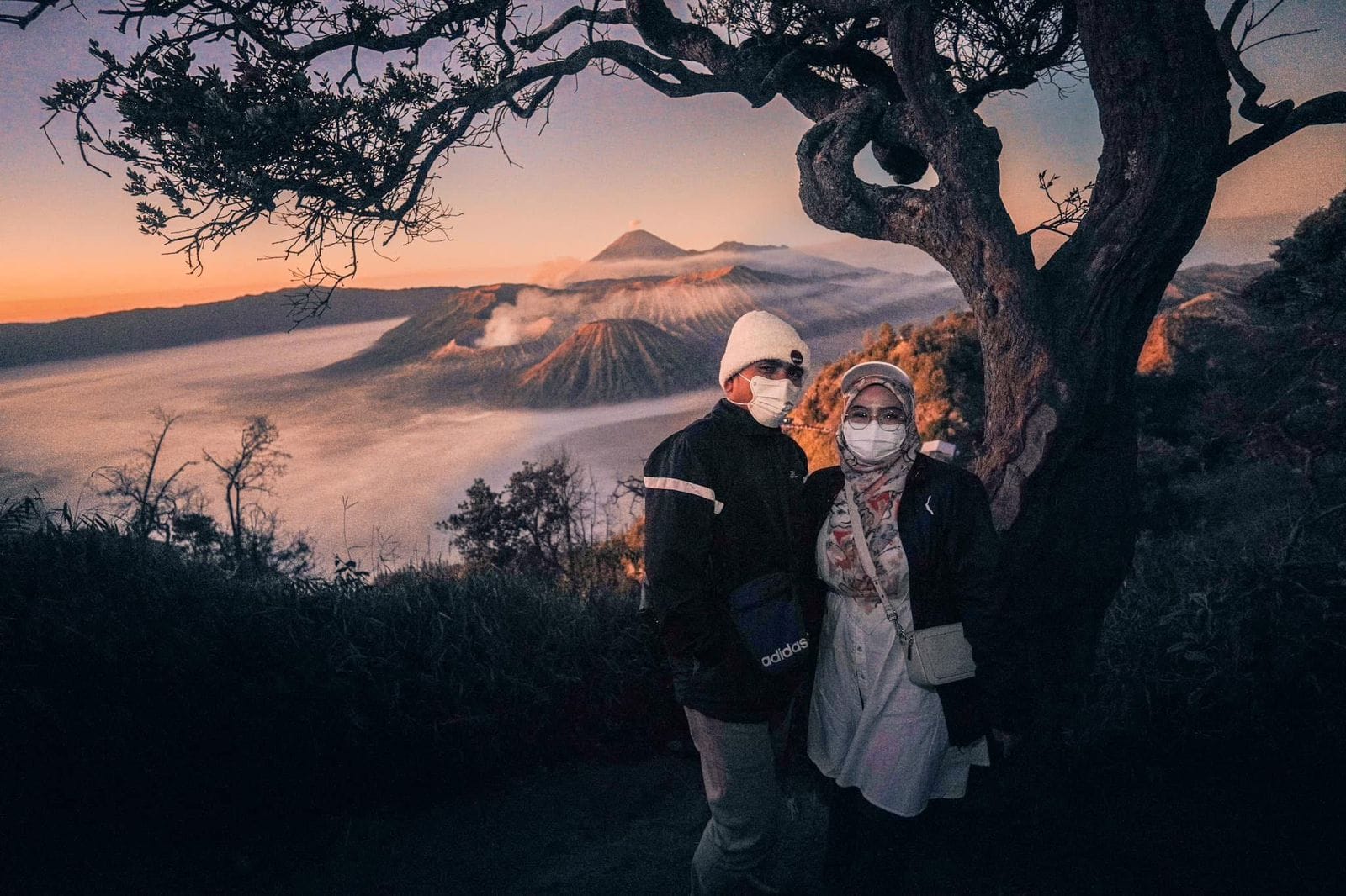Couple dressed in warm jackets, hats, and scarves posing at sunrise with Mount Bromo in the background, showing What to Wear for a Bromo Trip.