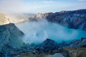 Aerial view of Mount Ijen crater with turquoise acid lake surrounded by volcanic rocks and mist at sunrise, captured through drone photography at Ijen sunrise showcasing the dramatic landscape and morning light.