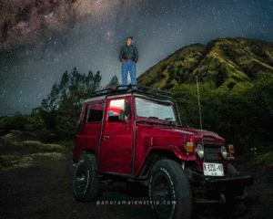 A traveler standing on top of a red Jeep Bromo under a stunning Milky Way night sky with the green hills of Mount Bromo in the background, capturing the adventurous spirit of exploring Bromo by jeep.