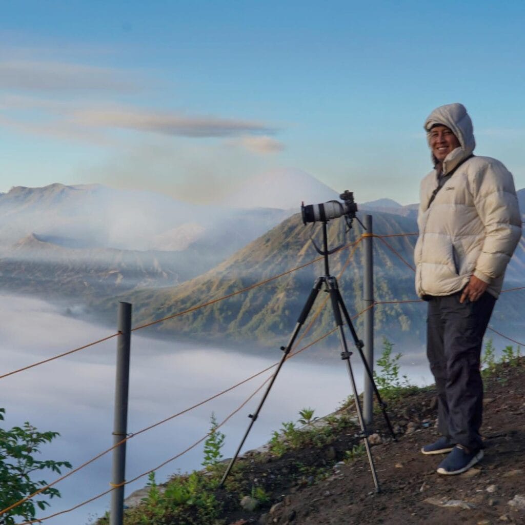 Photographer standing with camera tripod above misty mountains during Ijen sunrise & Blue Flame adventure in East Java.