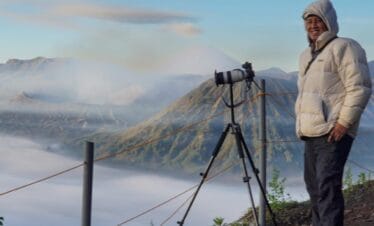 Photographer standing with camera tripod above misty mountains during Ijen sunrise & Blue Flame adventure in East Java.