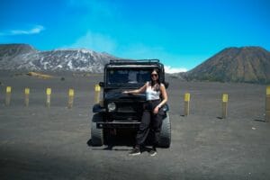 A traveler joyfully posing on a black Jeep with arms raised, surrounded by the dramatic volcanic terrain and bright blue sky, highlighting the breathtaking panorama Bromo during a sunny afternoon adventure.