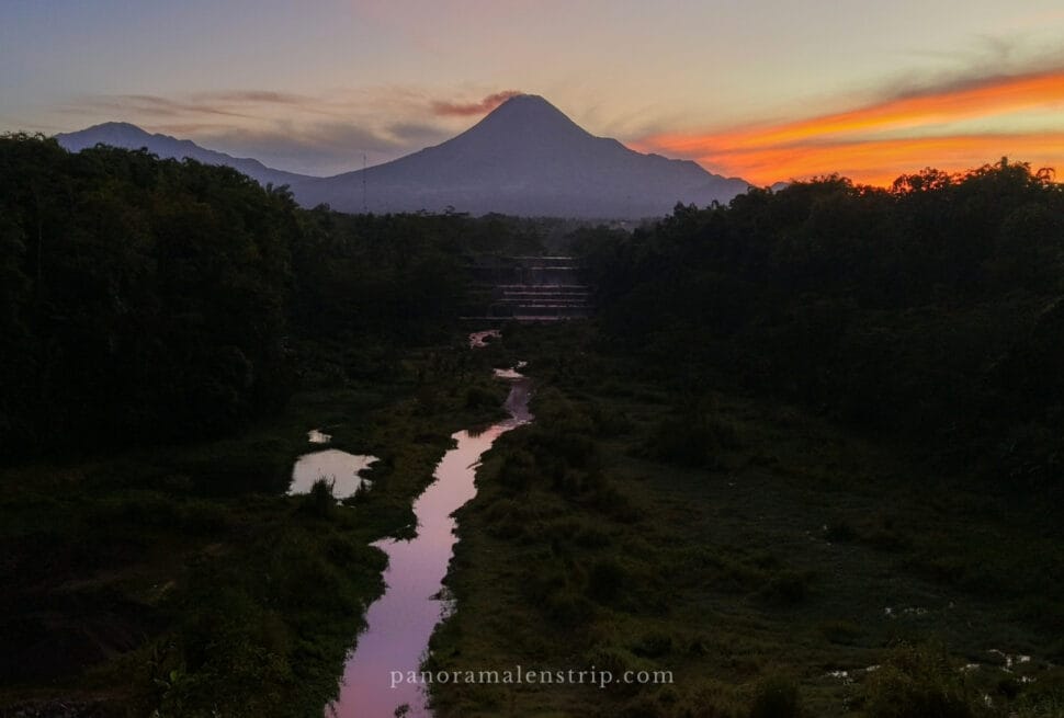 Sunrise view of Mount Merapi with glowing sky and a reflective river in the foreground.