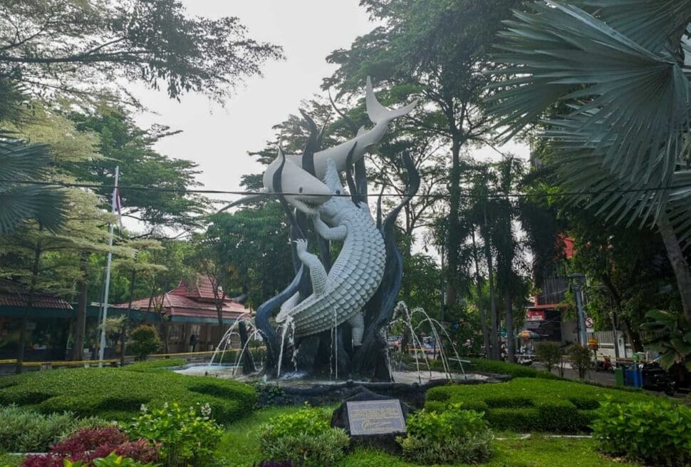 Iconic Suro and Boyo statue in Surabaya City Park, a popular landmark often visited during a Surabaya trip, surrounded by lush green trees and a small fountain.