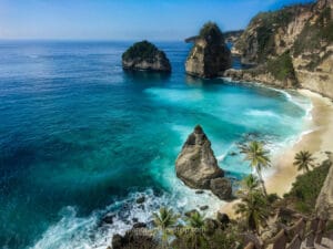 A stunning tropical beach view ideal for a Bali honeymoon, featuring turquoise waters, dramatic limestone cliffs, palm trees, and iconic rock formations along a serene coastline.