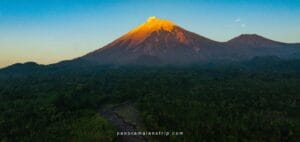 A majestic sunrise view of an active volcano during a Volcano Discovery journey, featuring a glowing volcanic peak above dense green forest and a winding river landscape.
