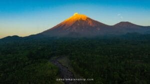 A majestic sunrise view of an active volcano during a Volcano Discovery journey, featuring a glowing volcanic peak above dense green forest and a winding river landscape.