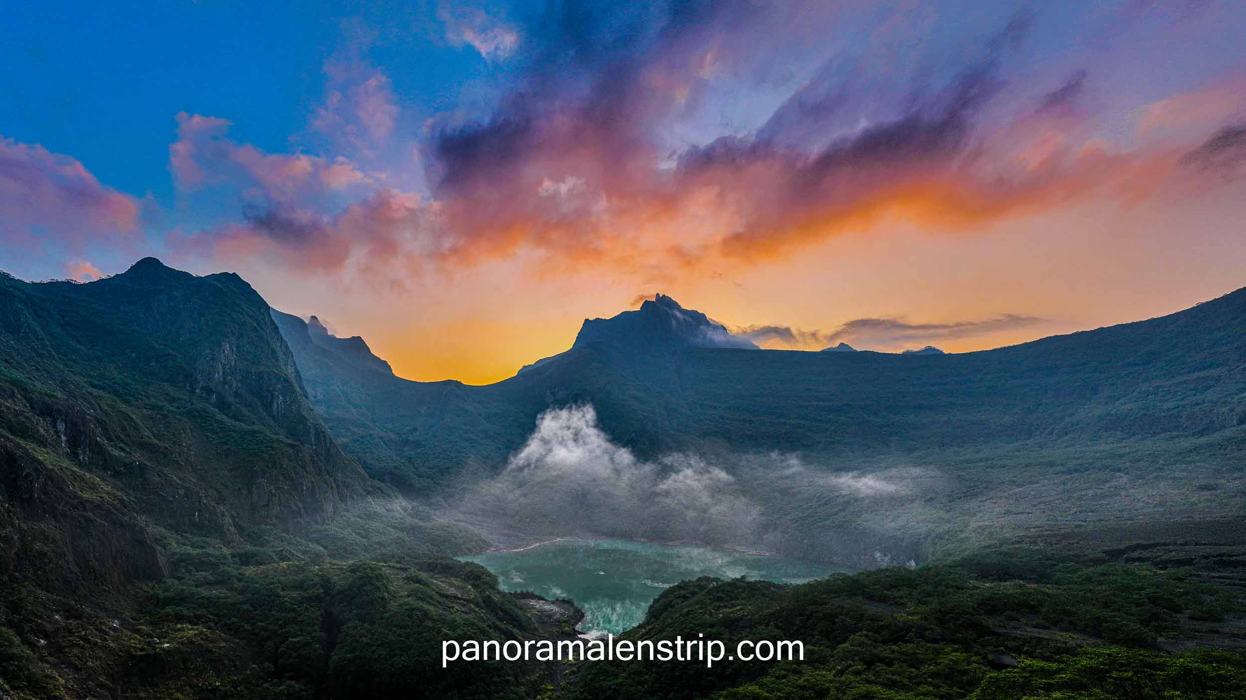 Sunrise view over the crater lake of Mount Kelud with dramatic volcanic cliffs, colorful sky, and mist rising from the lush green valley