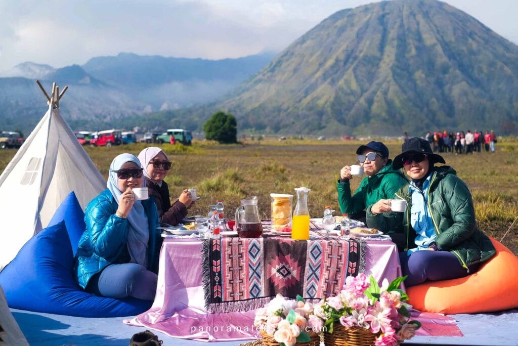 Picnic Bromo outdoor breakfast with women sitting around decorated picnic table, enjoying hot drinks with Mount Bromo and wide savanna background.