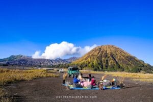 Picnic Bromo setup on the sea of sand with panoramic view of Mount Bromo, smoke from crater, green hills, and jeeps parked near the picnic area.