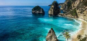 A stunning tropical beach view ideal for a Bali honeymoon, featuring turquoise waters, dramatic limestone cliffs, palm trees, and iconic rock formations along a serene coastline.