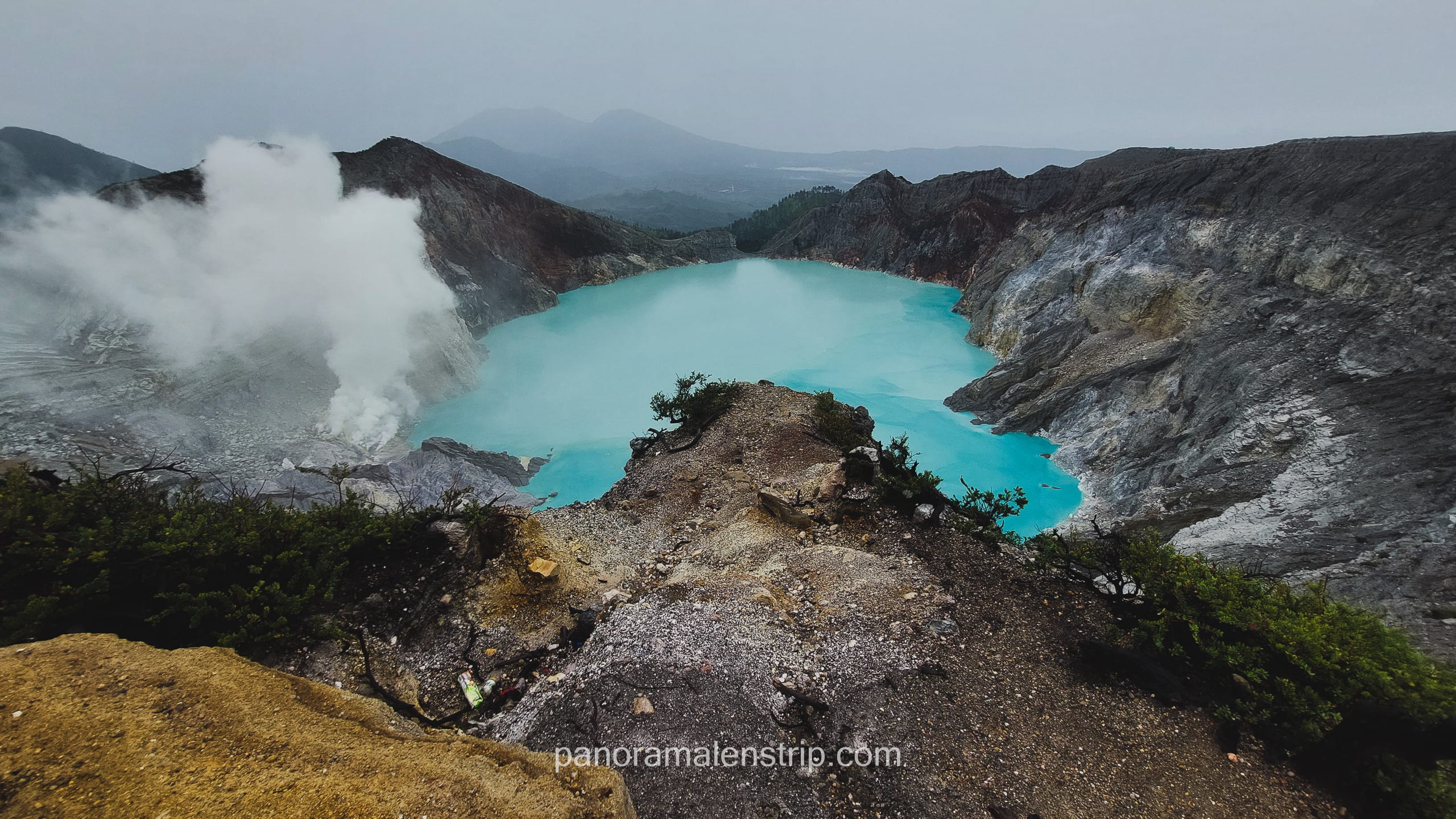 Panoramic view of a turquoise volcanic crater lake with steam rising from crater walls in a mountainous volcanic landscape