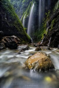 Long exposure photo of a tropical waterfall cascading down mossy green cliffs with flowing river and rocks in the foreground