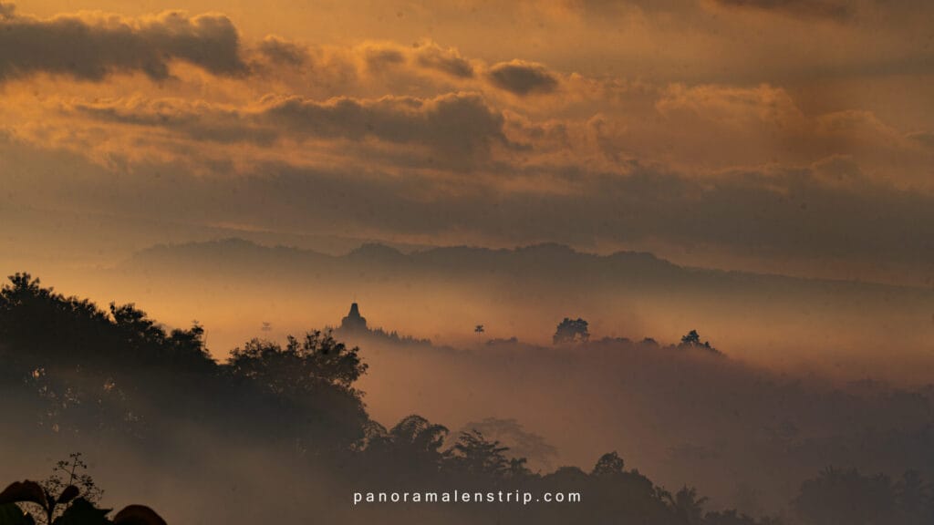 Sunrise view of the Borobudur UNESCO heritage site emerging from morning mist, surrounded by hills and dramatic clouds.