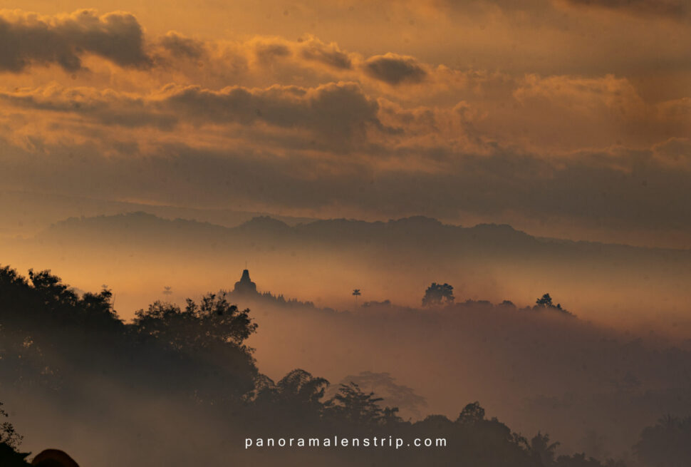Sunrise view of the Borobudur UNESCO heritage site emerging from morning mist, surrounded by hills and dramatic clouds.