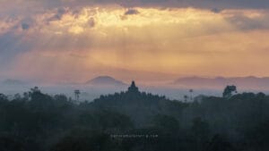 Borobudur UNESCO heritage site at sunrise, rising above misty forests with golden light and mountains in the background.