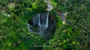 Looking up from the bottom of Tumpak Sewu Waterfall with misty water spray and dark volcanic rocks in the foreground.