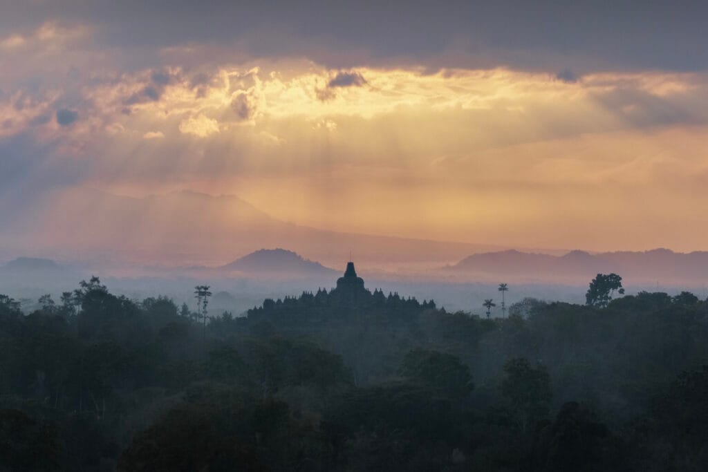 A dark silhouette of the Borobudur temple rising above a misty jungle in Indonesia, illuminated by golden sunbeams piercing through morning clouds.