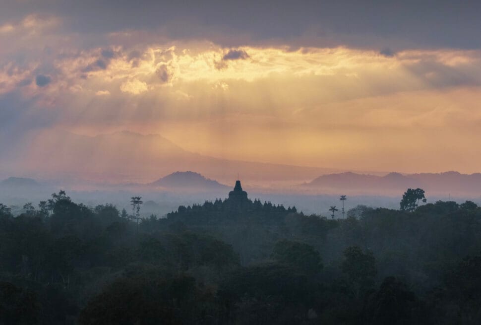 A dark silhouette of the Borobudur temple rising above a misty jungle in Indonesia, illuminated by golden sunbeams piercing through morning clouds.