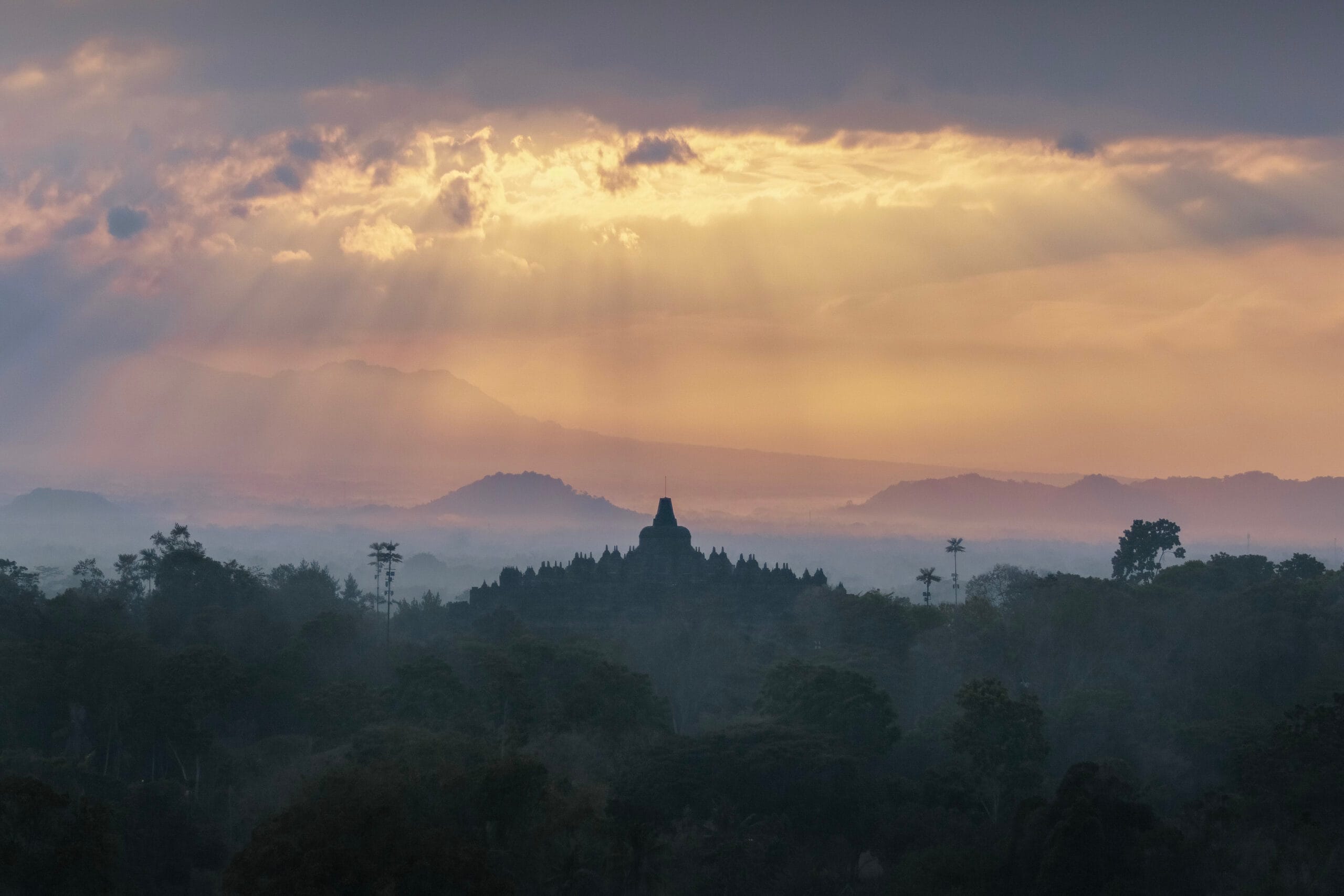 A dark silhouette of the Borobudur temple rising above a misty jungle in Indonesia, illuminated by golden sunbeams piercing through morning clouds.