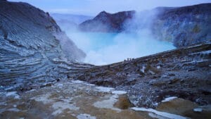 Close-up view during the Ijen Crater Hike showing steep volcanic rock formations, sulfur-stained crater walls, thick mist rising from the turquoise crater lake, and hikers standing along the rim at Mount Ijen, East Java, Indonesia.