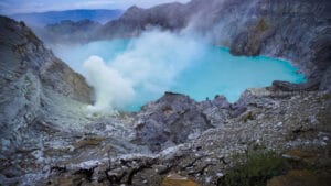 Scenic view during the Ijen Crater Hike highlighting the vivid turquoise acidic lake, thick sulfur smoke rising from volcanic vents, steep rocky crater walls, and hikers descending toward the crater floor at Mount Ijen, East Java, Indonesia.