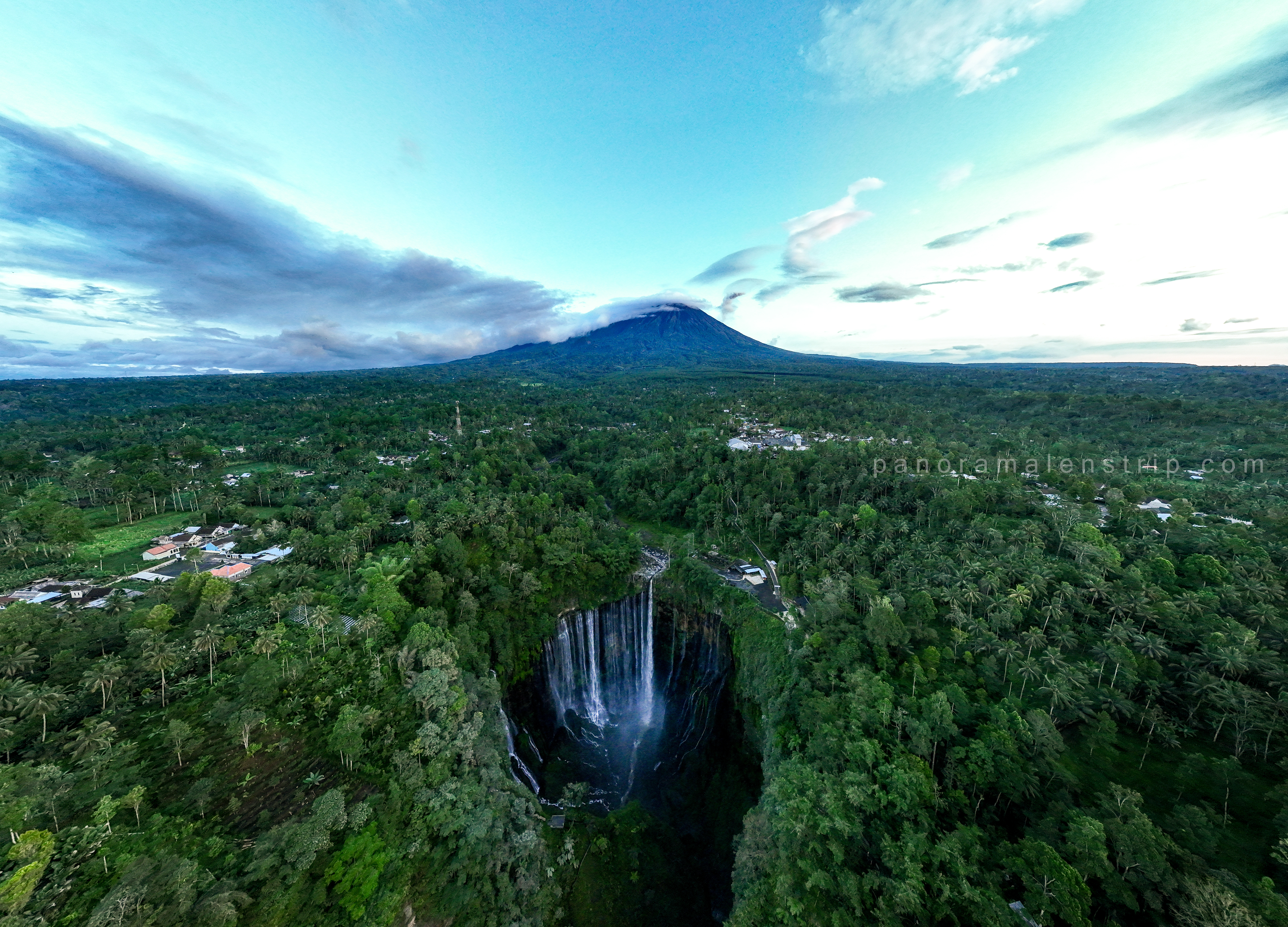 Tumpak Sewu photography capturing a dramatic panoramic view of the multi-tiered waterfall surrounded by lush tropical rainforest in East Java, Indonesia, with misty cascades flowing down steep green cliffs under a bright sky.