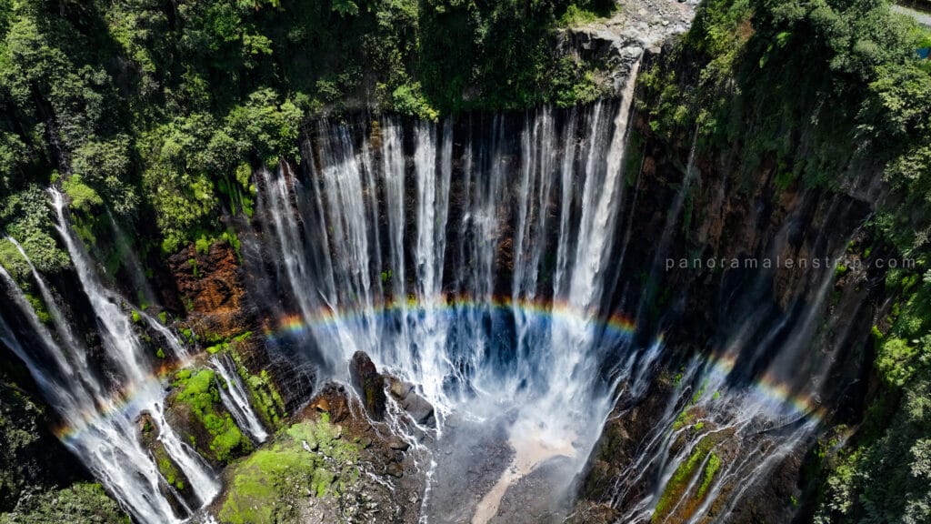 Tumpak Sewu photography showcasing a dramatic aerial view of the Tumpak Sewu Waterfall in East Java, Indonesia, with multiple cascading streams plunging into a lush green canyon and a visible rainbow forming in the mist below.