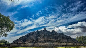 Borobudur temple climb view from ground level showing the massive stone terraces under dramatic blue sky in Central Java, Indonesia.