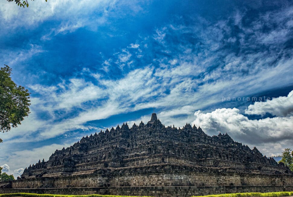 Borobudur temple climb view from ground level showing the massive stone terraces under dramatic blue sky in Central Java, Indonesia.