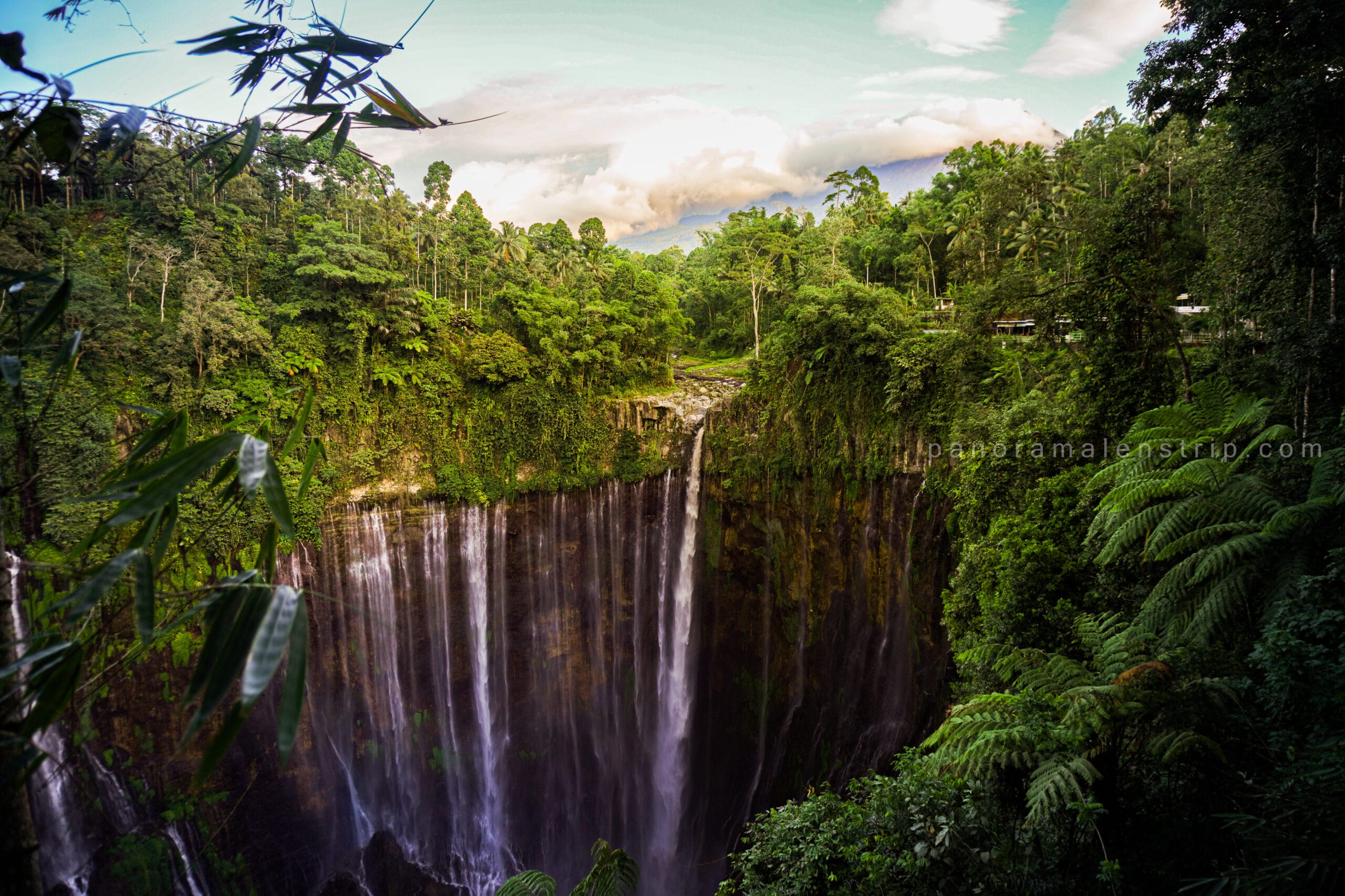 Tumpak Sewu photography capturing a dramatic panoramic view of the multi-tiered waterfall surrounded by lush tropical rainforest in East Java, Indonesia, with misty cascades flowing down steep green cliffs under a bright sky.