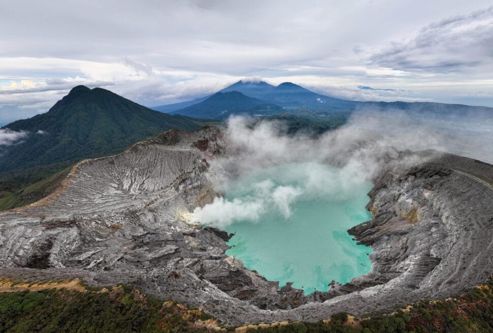 High-angle panoramic view of Mount Ijen's volcanic crater featuring the turquoise lake and billowing sulfur smoke under a cloudy sky.