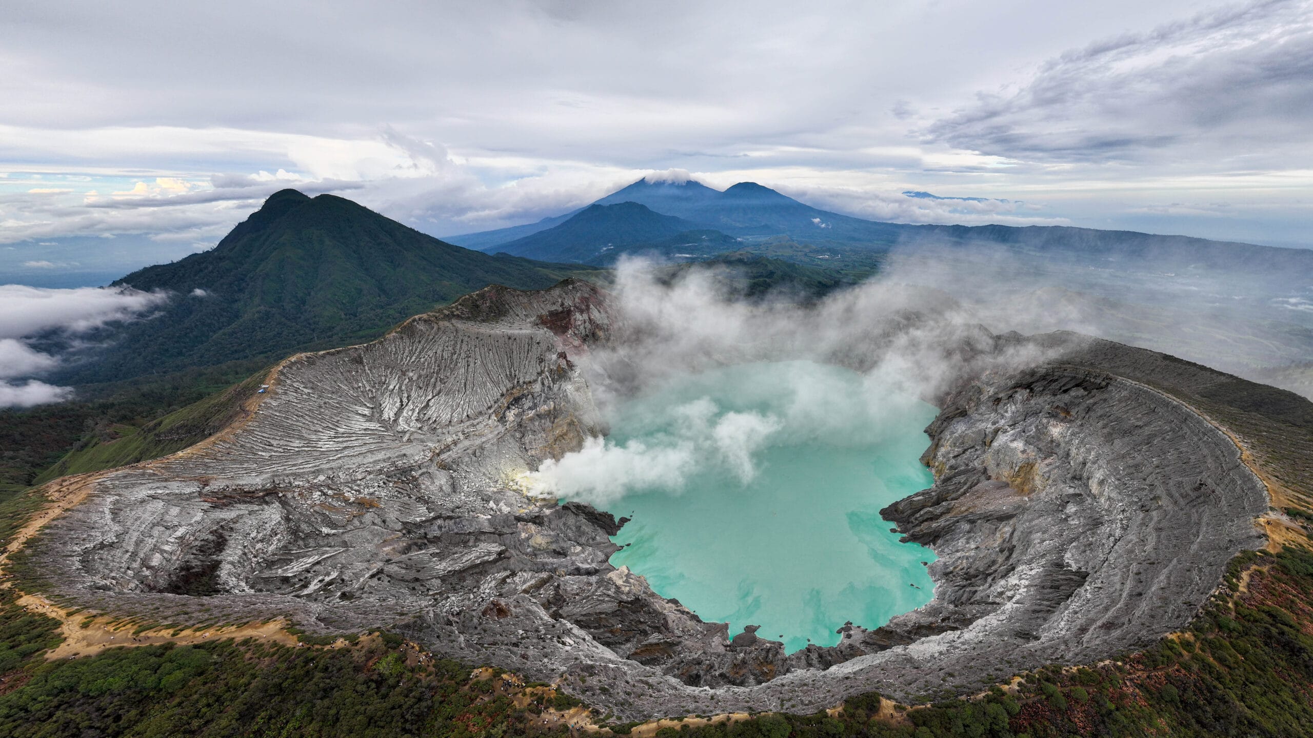 High-angle panoramic view of Mount Ijen's volcanic crater featuring the turquoise lake and billowing sulfur smoke under a cloudy sky.