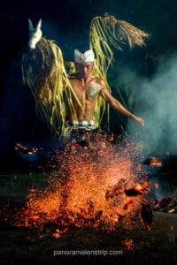 A shirtless man wearing a traditional Balinese udeng and checkered sarong kicks burning embers, creating a dramatic burst of orange sparks in the dark. He carries a woven palm-leaf prop featuring a white horse mask on his shoulders during a fire dance.