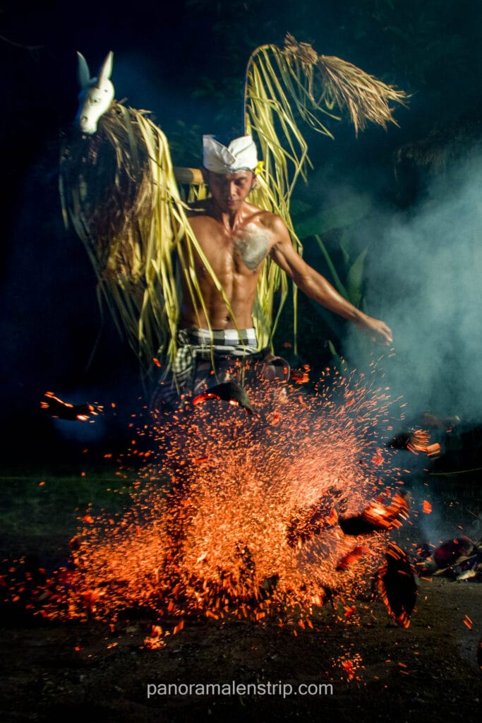 A shirtless man wearing a traditional Balinese udeng and checkered sarong kicks burning embers, creating a dramatic burst of orange sparks in the dark. He carries a woven palm-leaf prop featuring a white horse mask on his shoulders during a fire dance.