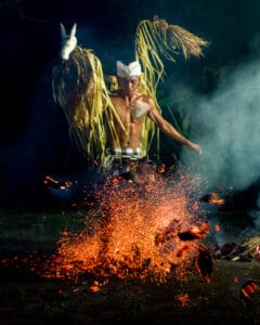 A shirtless man wearing a traditional Balinese udeng and checkered sarong kicks burning embers, creating a dramatic burst of orange sparks in the dark. He carries a woven palm-leaf prop featuring a white horse mask on his shoulders during a fire dance.