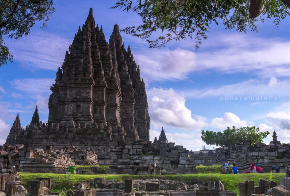 The towering stone spires of the 9th-century Prambanan Hindu Temple complex in Yogyakarta, Indonesia, under a blue sky with scattered clouds and lush green grass in the foreground.