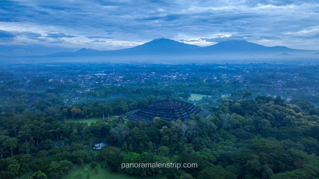 Aerial drone view of the Borobudur Temple pyramid surrounded by mist and forest with Mount Merapi in the background at dawn.