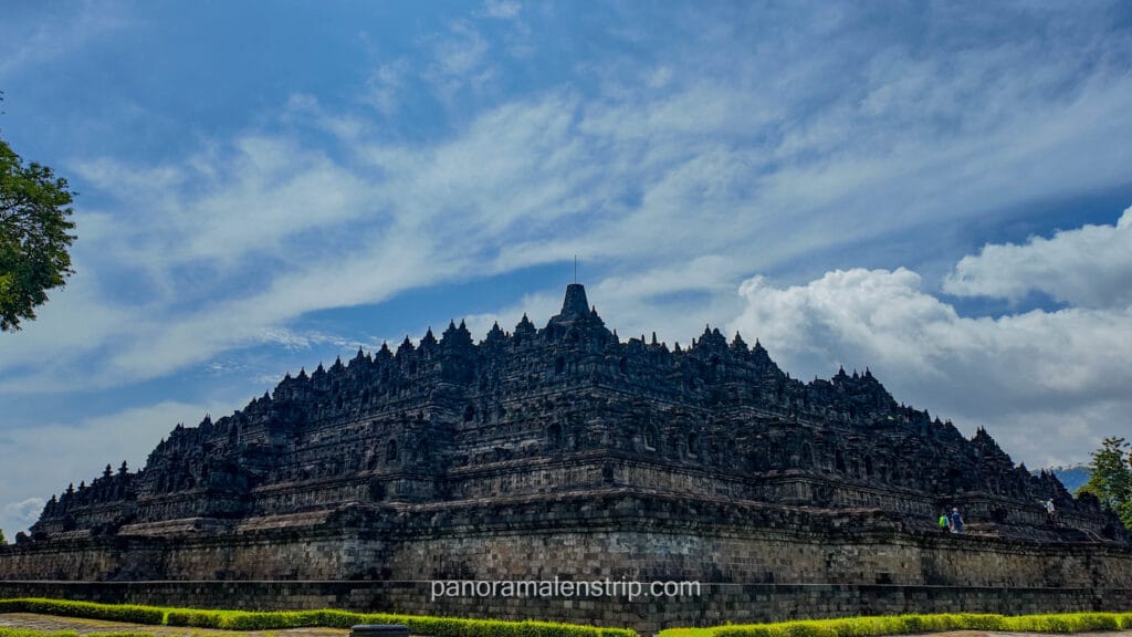 A low-angle full view of the massive tiered pyramid structure of Borobudur Temple against a blue sky with clouds.