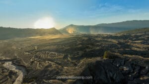 A wide panoramic view of the Mount Bromo volcanic caldera at sunrise with golden light hitting the dusty sea of sand and mountain ridges.