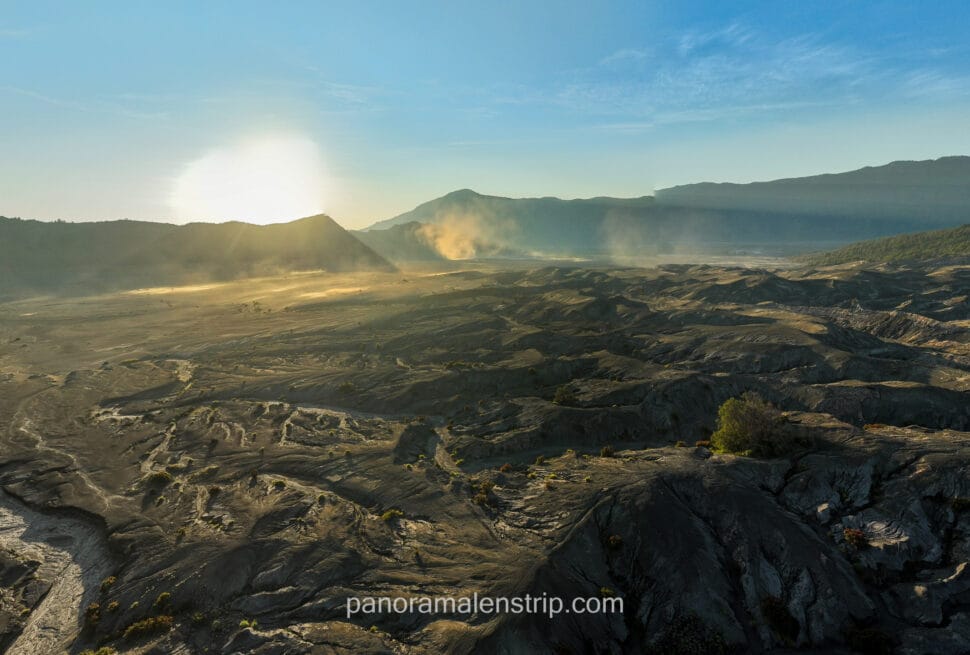 A wide panoramic view of the Mount Bromo volcanic caldera at sunrise with golden light hitting the dusty sea of sand and mountain ridges.