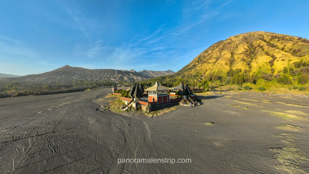 The Pura Luhur Poten Hindu temple sitting in the middle of the Bromo Sea of Sand with a lush green mountain in the background under a blue sky.