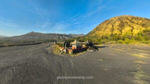 The Pura Luhur Poten Hindu temple sitting in the middle of the Bromo Sea of Sand with a lush green mountain in the background under a blue sky.