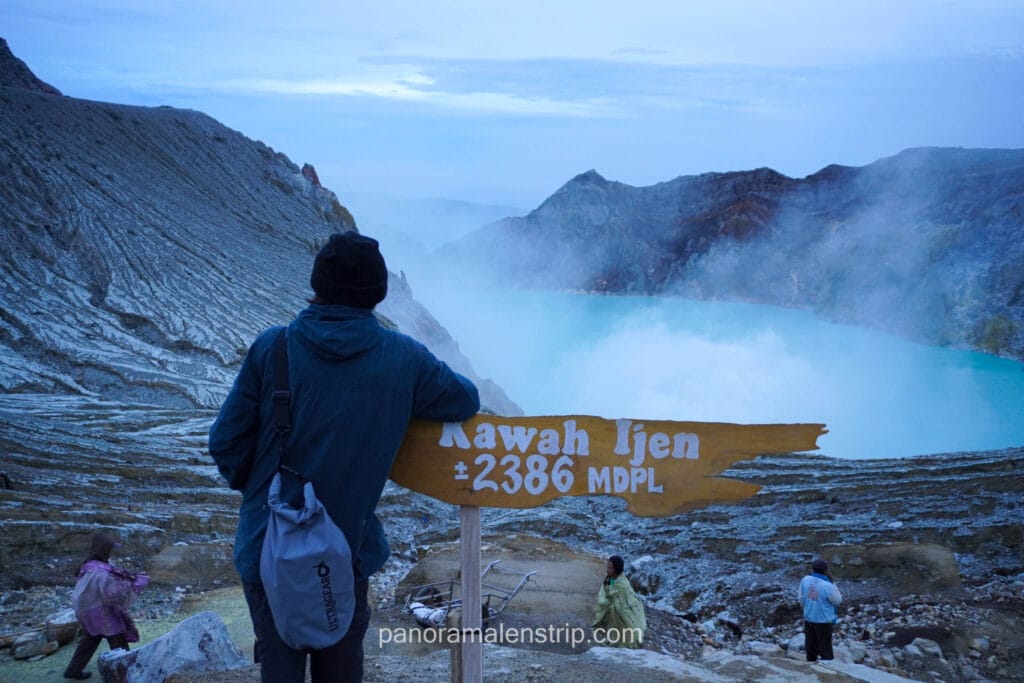 A hiker standing next to the wooden Kawah Ijen elevation sign at 2386 MDPL overlooking the turquoise crater lake.