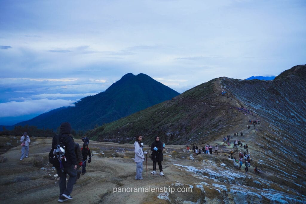 A group of hikers walking along the Kawah Ijen crater rim trail at dawn with a view of neighboring volcanic peaks.