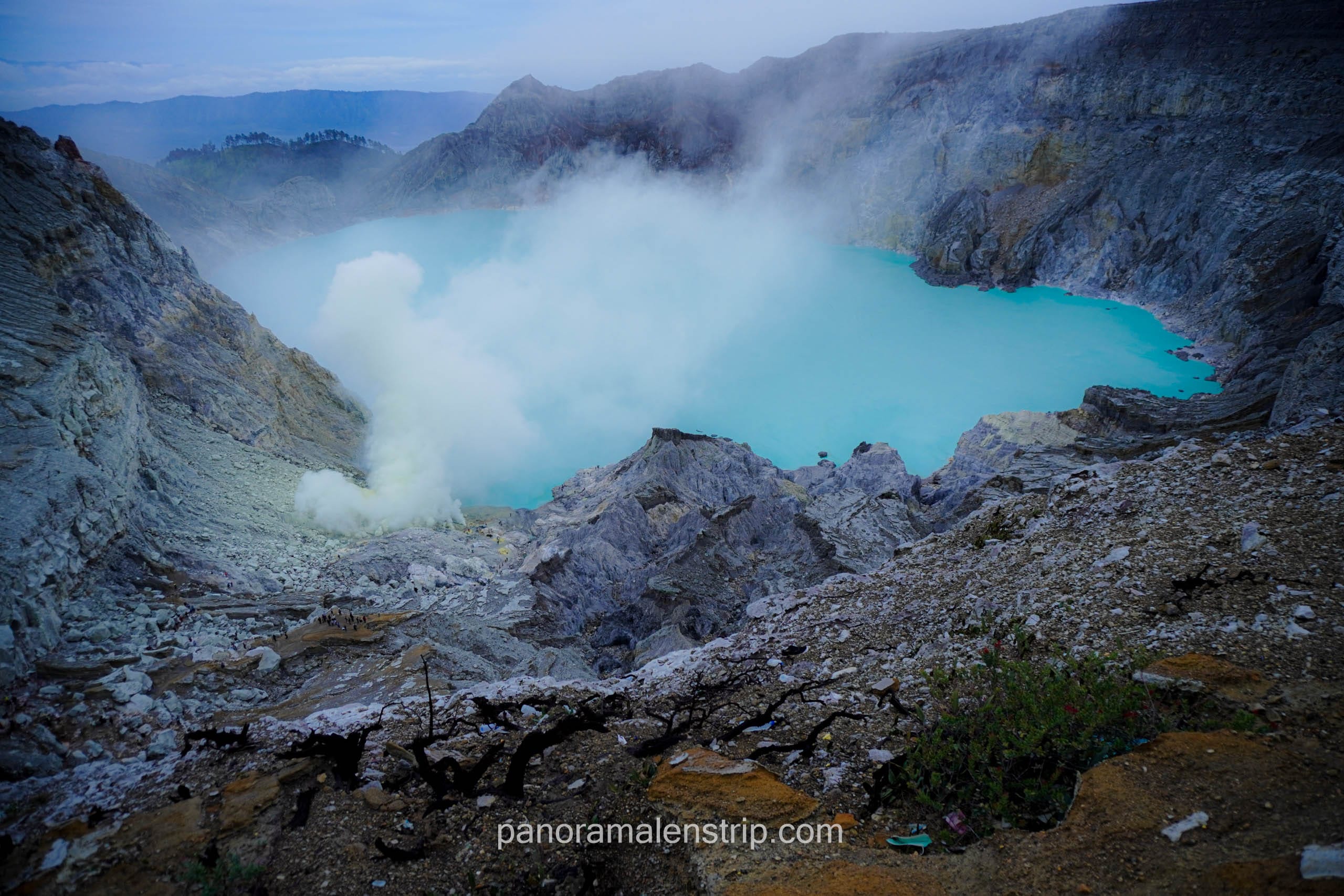 Panoramic landscape of the Kawah Ijen turquoise acid lake with thick white sulfur smoke rising from the active vents.