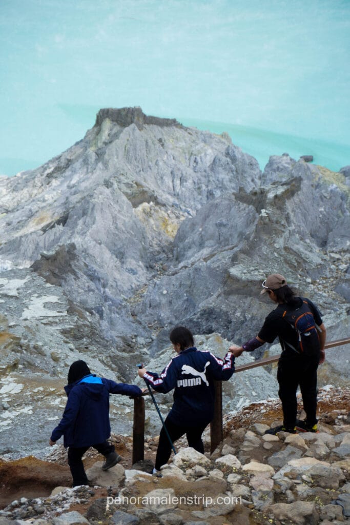 Tourists and a guide navigating the steep, rocky hiking trail descending into the Ijen Crater with the turquoise lake in the background.